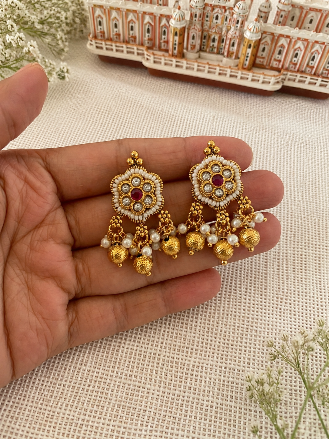 Gold and pearl earrings held in a hand with a decorative building in the background