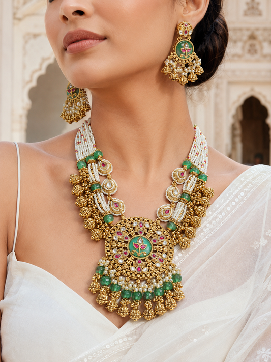 Woman wearing a gold necklace and earrings with green and red accents in an outdoor setting.
