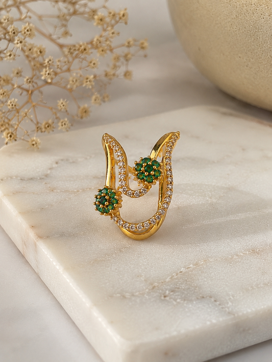 A close-up view of a gold-toned Vanki chevron ring adorned with pave clear CZ crystals and two emerald-green floral stone clusters resting on marble.