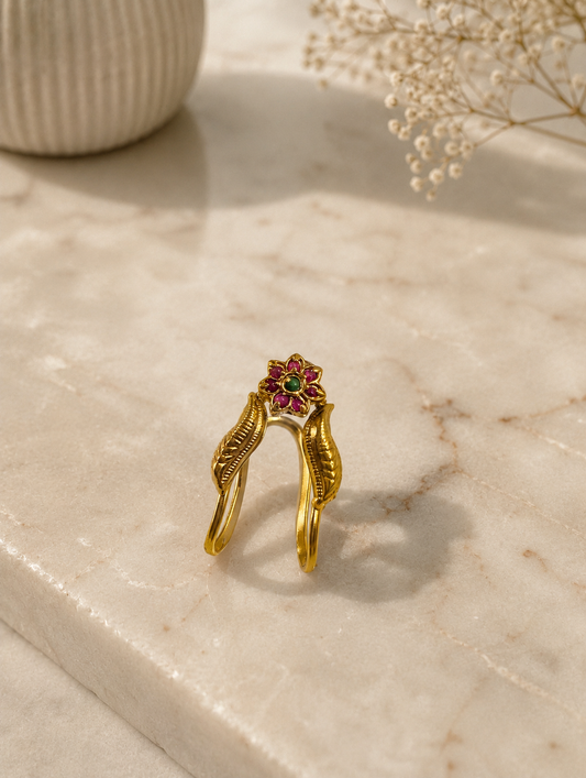 An antique gold-toned Vanki ring resting upright on a marble surface, featuring carved leaf details along its V-curved band and a central floral cluster encrusted with ruby-red stones and a green stone center.