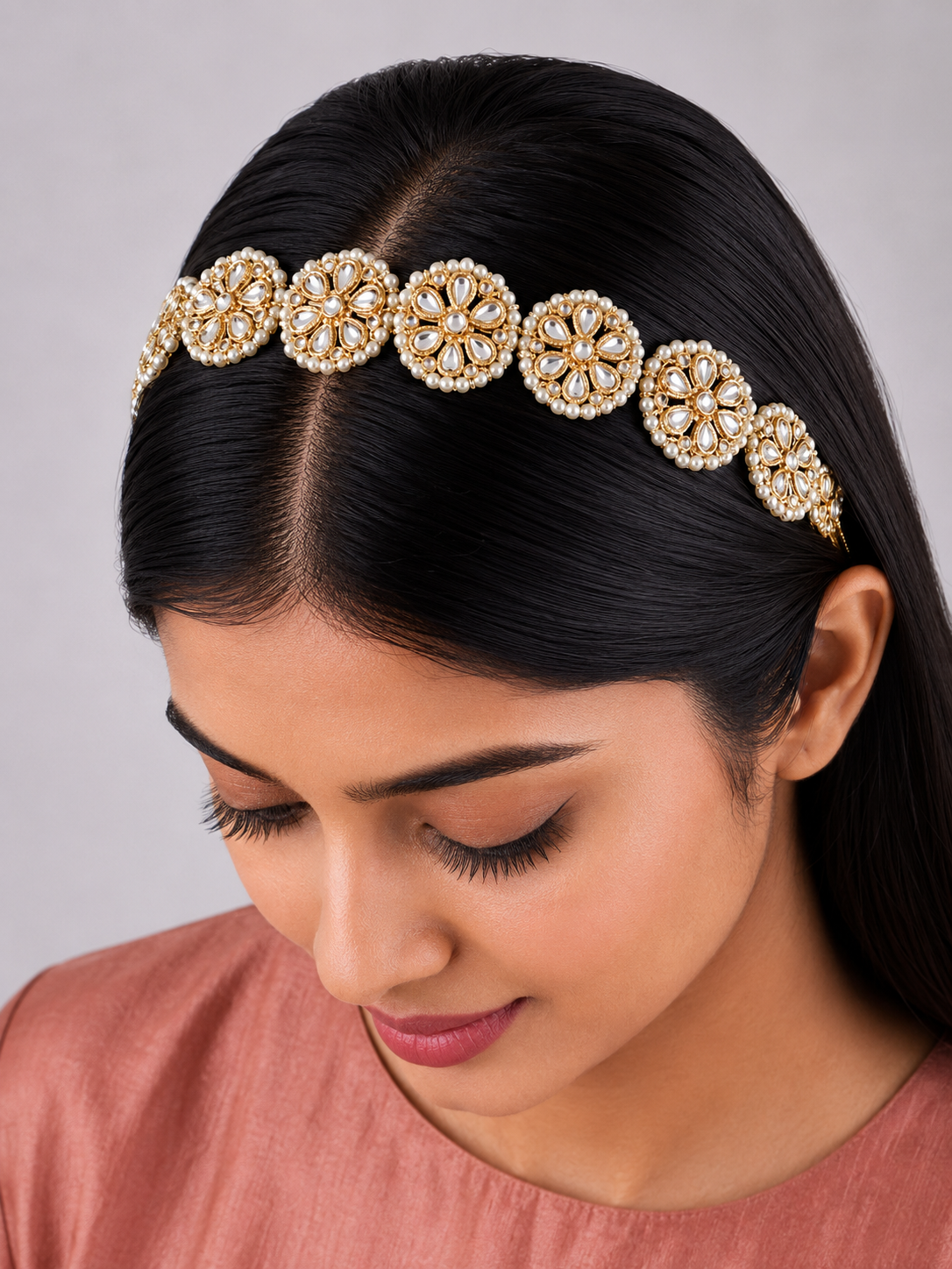 Traditional Indian gold-plated sheeshphool hair band featuring a continuous row of circular floral motifs set with glistening Kundan stones and tiny white pearls, shown laid flat on rich maroon satin fabric and modeled on a woman's hair.