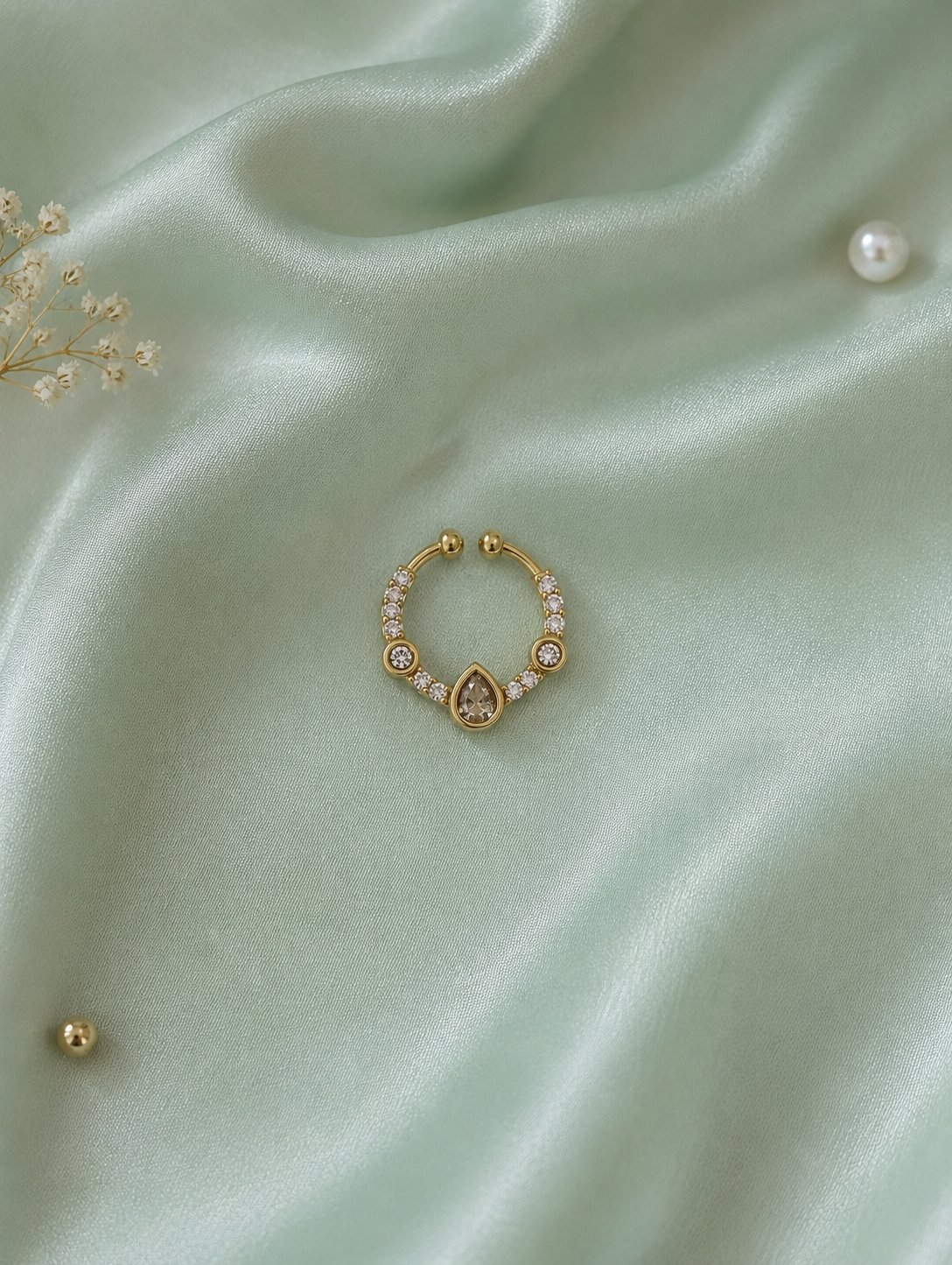 Elegant gold-toned septum ring featuring a central teardrop crystal and surrounding white diamond accents, displayed on a textured sage green silk background.
