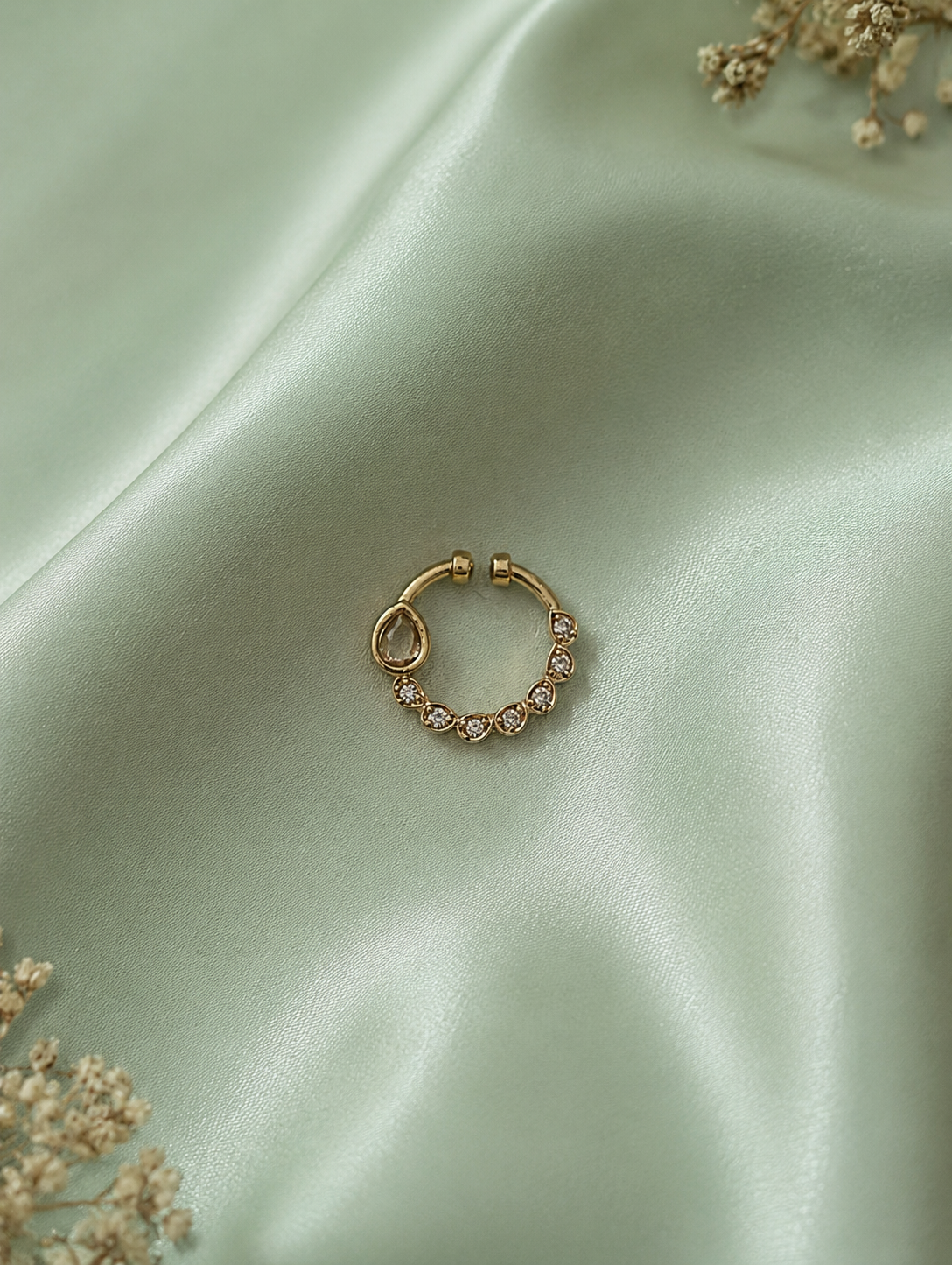 An asymmetric gold-toned septum ring with an off-center teardrop crystal and a curved row of small white diamonds, resting on a draped mint green silk background with faint dried floral accents.