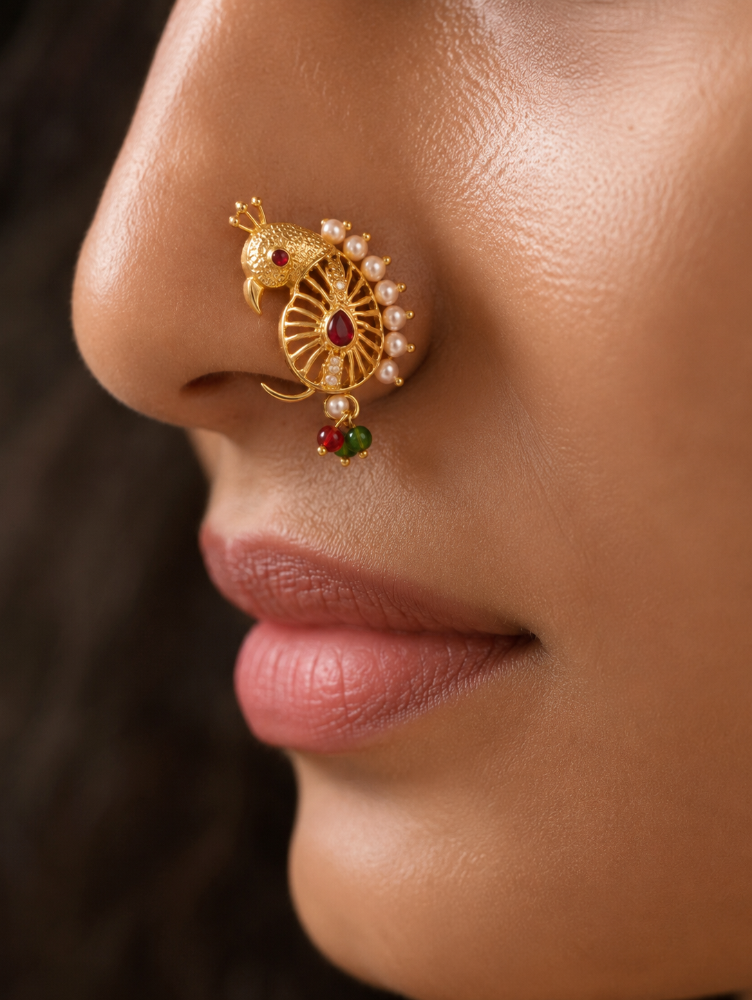 Close-up of a model wearing a traditional gold Maharashtrian Peshwai Nath nose ring featuring an intricate peacock design accented with a ruby-red gemstone, delicate white pearls, and tiny hanging red and green beads.