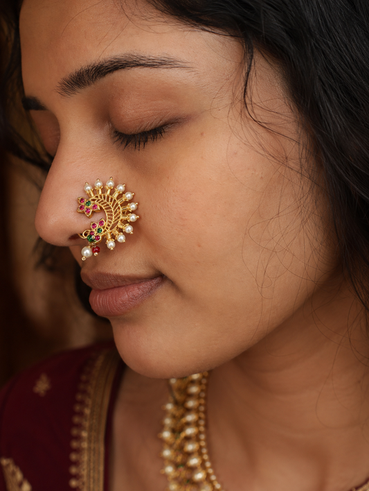 A close-up of a woman wearing a gold crescent-shaped nose ornament (nose stud) decorated with small white pearls and red and green stones, paired with a gold necklace.
