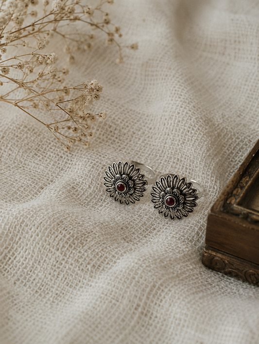 Silver sunflower-shaped earrings with red stones on a textured beige fabric background.