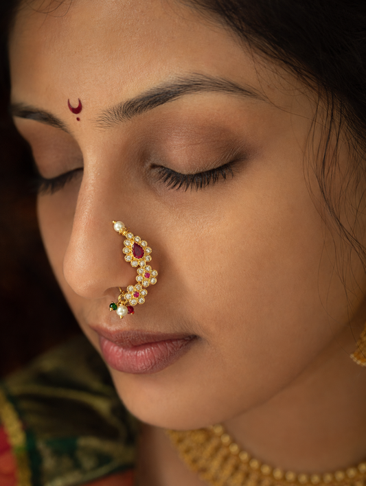 A close-up of a woman with her eyes closed, wearing a gold crescent moon nose ornament (nath) adorned with white pearls and a red gemstone, along with a red bindi on her forehead.