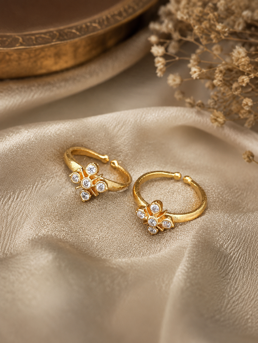 A pair of gold hoop earrings with small flower-like clusters of clear stones, displayed on draped beige fabric with dried flowers and a gold-toned bowl in the background.