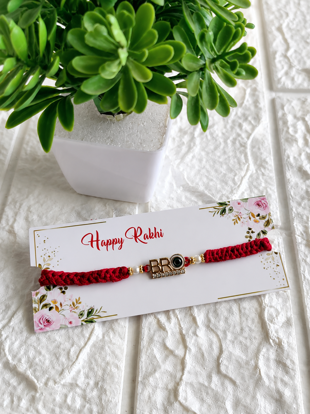 A modern Raksha Bandhan Rakhi featuring a thick red braided cord and a golden centerpiece that spells "BRO" with rhinestone accents, flanked by small pearls, displayed on a white decorative card with "Happy Rakhi" script alongside a small green potted plant.