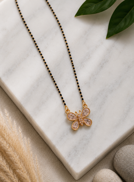 A product photography showcase of a modern gold-toned mangalsutra featuring a butterfly pendant set with clear crystals and a traditional fine chain interspersed with dainty black beads. The collection includes a styled flat-lay layout on a white marble slab surrounded by textured textiles and botanical accents, and a macro close-up view detailing the stone-encrusted butterfly charm.