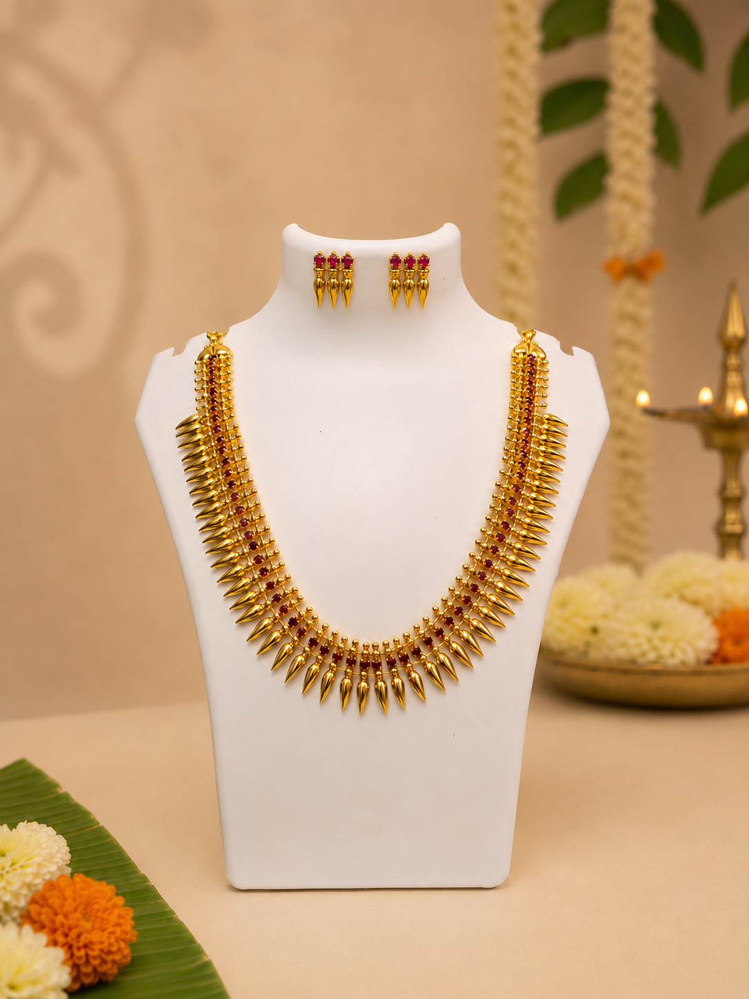 A traditional gold-plated Kerala Mullu Mottu Haram necklace set featuring jasmine bud spike motifs with small ruby red accents, displayed alongside matching stud earrings on a white jewelry bust in a festive setting.