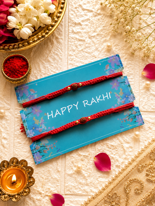 Set of two deep red braided Rakhi threads featuring a central natural Rudraksha bead flanked by gold accent beads on a blue Happy Rakhi card.