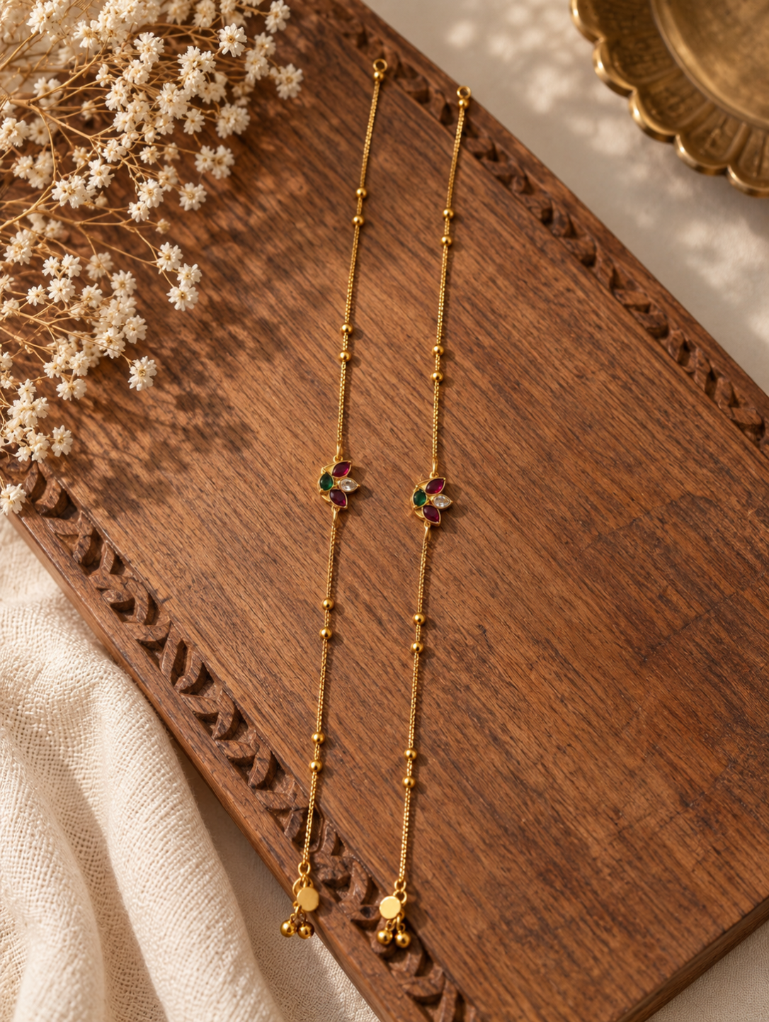 Pair of delicate gold bead anklets featuring ruby and emerald stone floral charms displayed on a carved wooden surface.