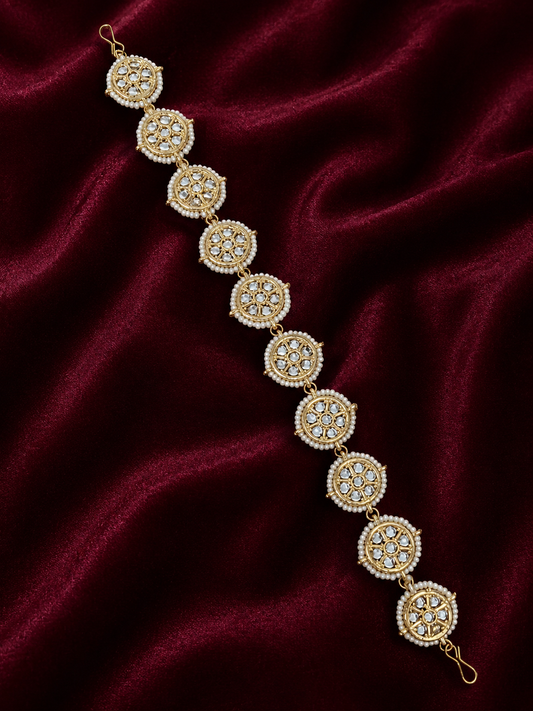 Traditional gold-plated Indian sheeshphool hair accessory featuring a flexible row of circular wheel motifs studded with Kundan stones and framed by tiny white pearls, elegantly displayed against a rich burgundy velvet backdrop.