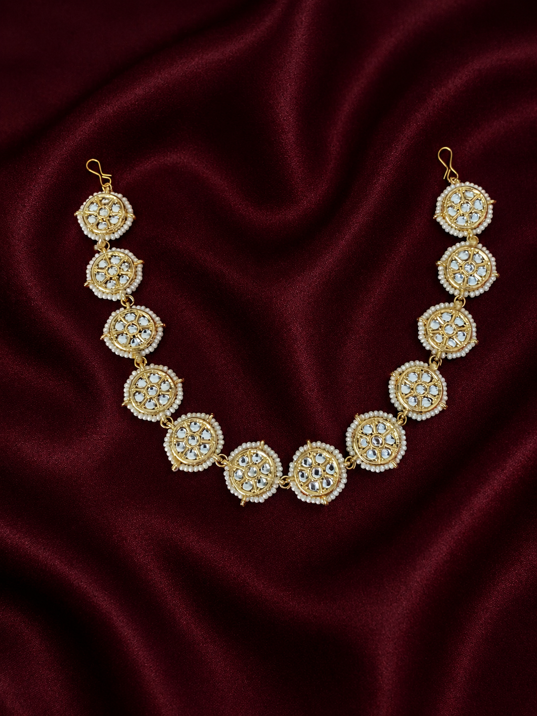 Traditional gold-plated Indian sheeshphool hair accessory featuring a flexible row of circular wheel motifs studded with Kundan stones and framed by tiny white pearls, elegantly displayed against a rich burgundy velvet backdrop.