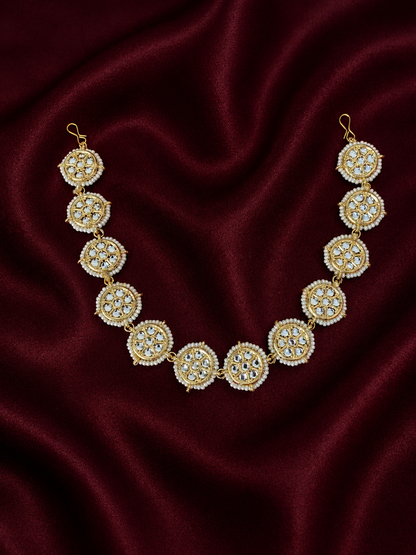 Traditional gold-plated Indian sheeshphool hair accessory featuring a flexible row of circular wheel motifs studded with Kundan stones and framed by tiny white pearls, elegantly displayed against a rich burgundy velvet backdrop.