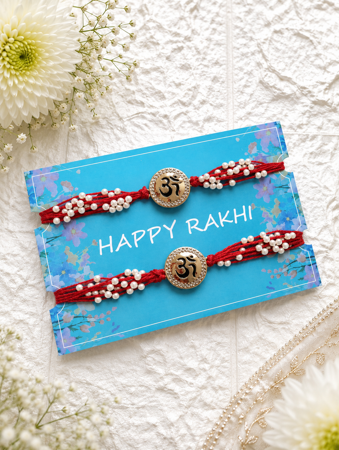 A set of two matching red thread Rakhis featuring gold Om centerpieces embellished with small pearls, mounted on a blue "Happy Rakhi" card.