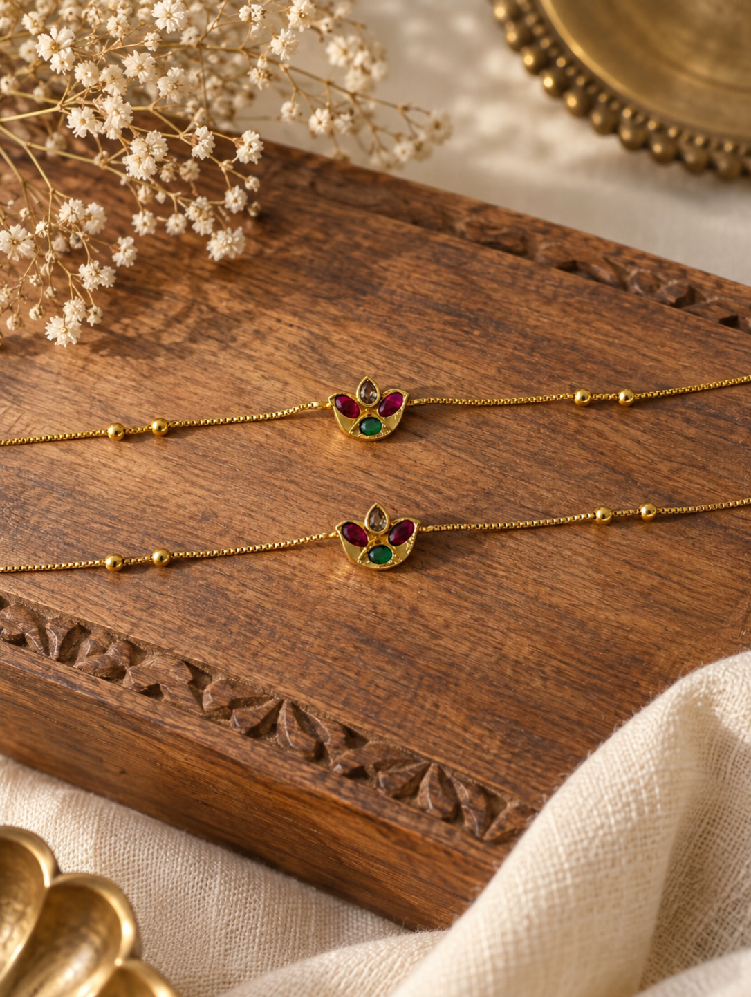 Pair of delicate gold bead anklets featuring ruby and emerald stone floral charms displayed on a carved wooden surface.