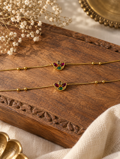 Pair of delicate gold bead anklets featuring ruby and emerald stone floral charms displayed on a carved wooden surface.