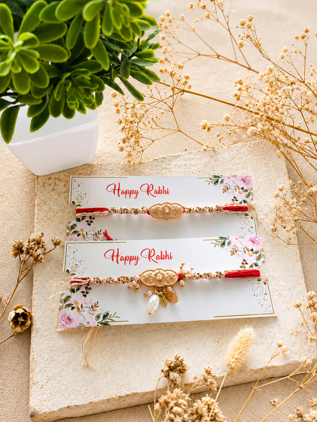 Two decorative Rakhi bracelets displayed on white floral cards that read 'Happy Rabbi,' arranged on a beige stone surface with dried flowers and a green plant in the background.