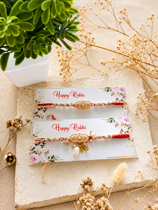 Two decorative Rakhi bracelets displayed on white floral cards that read 'Happy Rabbi,' arranged on a beige stone surface with dried flowers and a green plant in the background.