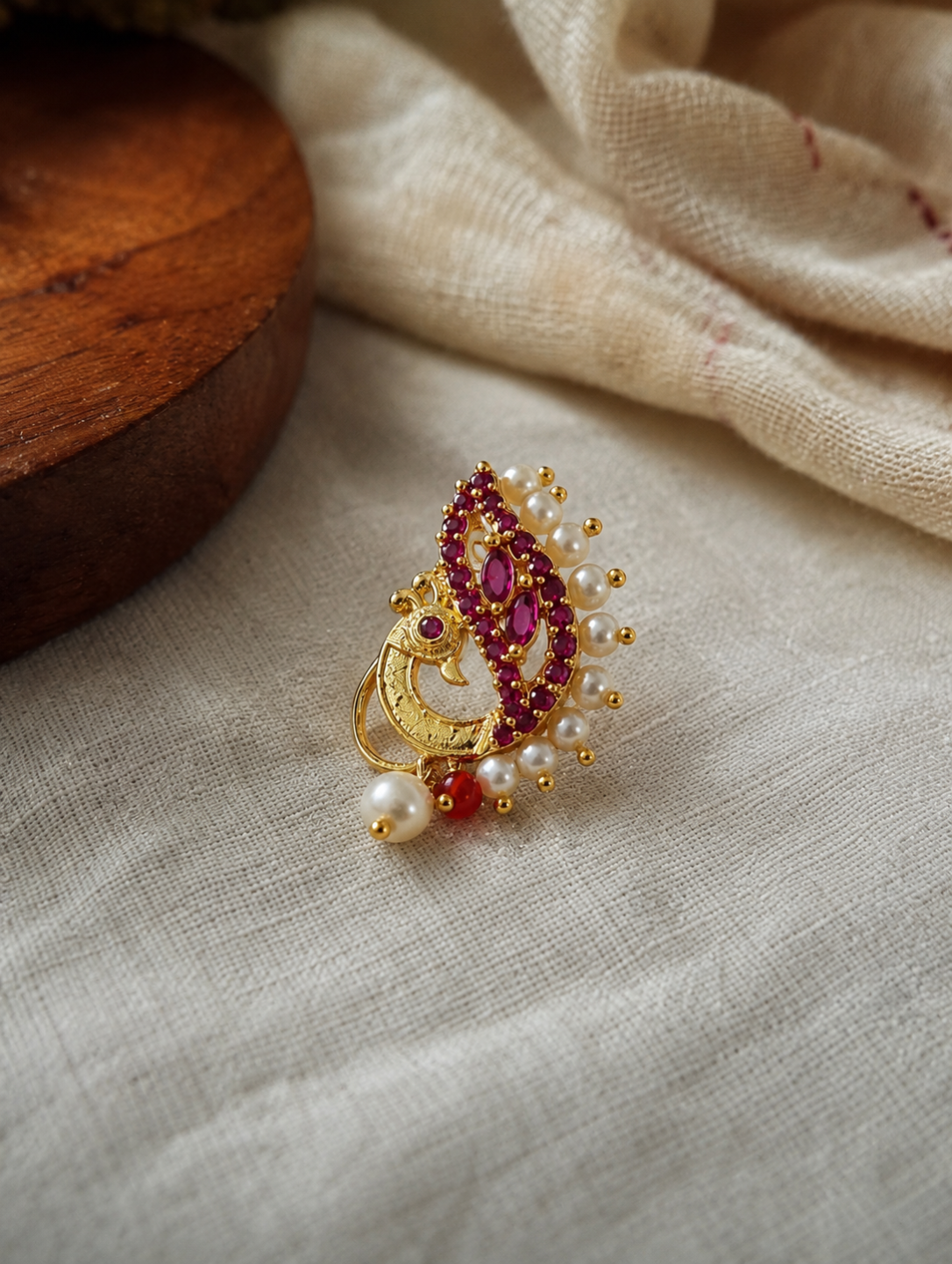 A gold-toned traditional Indian brooch featuring a peacock motif, embellished with deep pinkish-red stones, white faux pearls, and small gold beads, resting on a textured beige linen fabric next to a wooden block.