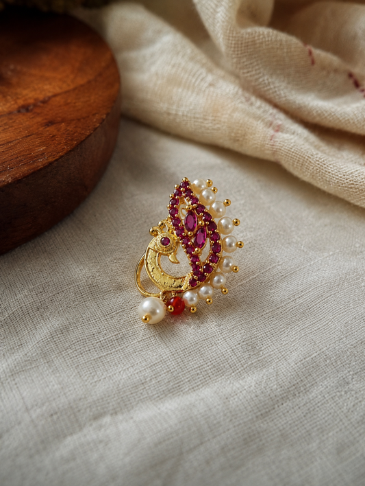 A gold-toned traditional Indian brooch featuring a peacock motif, embellished with deep pinkish-red stones, white faux pearls, and small gold beads, resting on a textured beige linen fabric next to a wooden block.