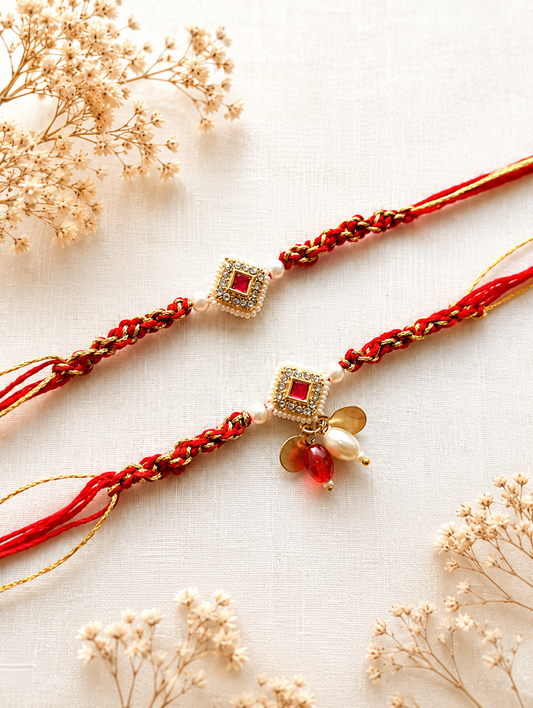 Two handcrafted designer Rakhis featuring square gold Kundan centers with ruby red gemstones, white pearl charms, and thick braided red and gold threads.