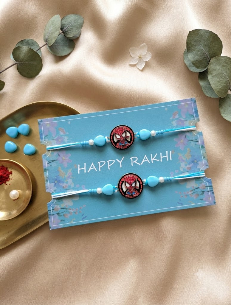 Two handmade Spiderman Rakhis for kids on a blue card with "HAPPY RAKHI" text and floral patterns. The card is placed on a shiny beige satin fabric next to eucalyptus leaves, a small gold dish with roli tikka and rice, and loose blue heart-shaped beads. The perspective is top-down.