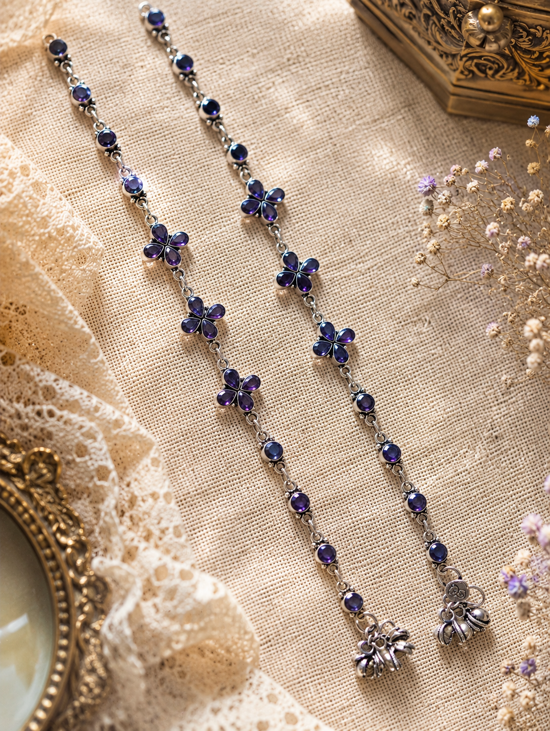 An overhead flat-lay view of a pair of handcrafted silver anklets from the Shika collection, laid out vertically on a cream textured canvas backdrop. The anklets feature deep purple stones in four-petal floral motifs and round links, styled alongside white lace trim, dried baby's breath flowers, and a vintage gold frame.