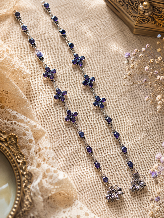 An overhead flat-lay view of a pair of handcrafted silver anklets from the Shika collection, laid out vertically on a cream textured canvas backdrop. The anklets feature deep purple stones in four-petal floral motifs and round links, styled alongside white lace trim, dried baby's breath flowers, and a vintage gold frame.
