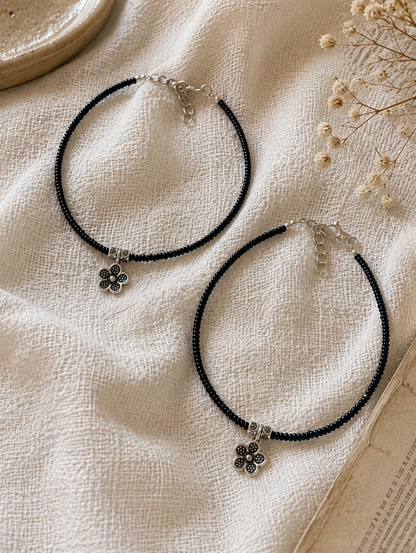 Two black beaded anklets with silver flower pendants on a textured beige surface.