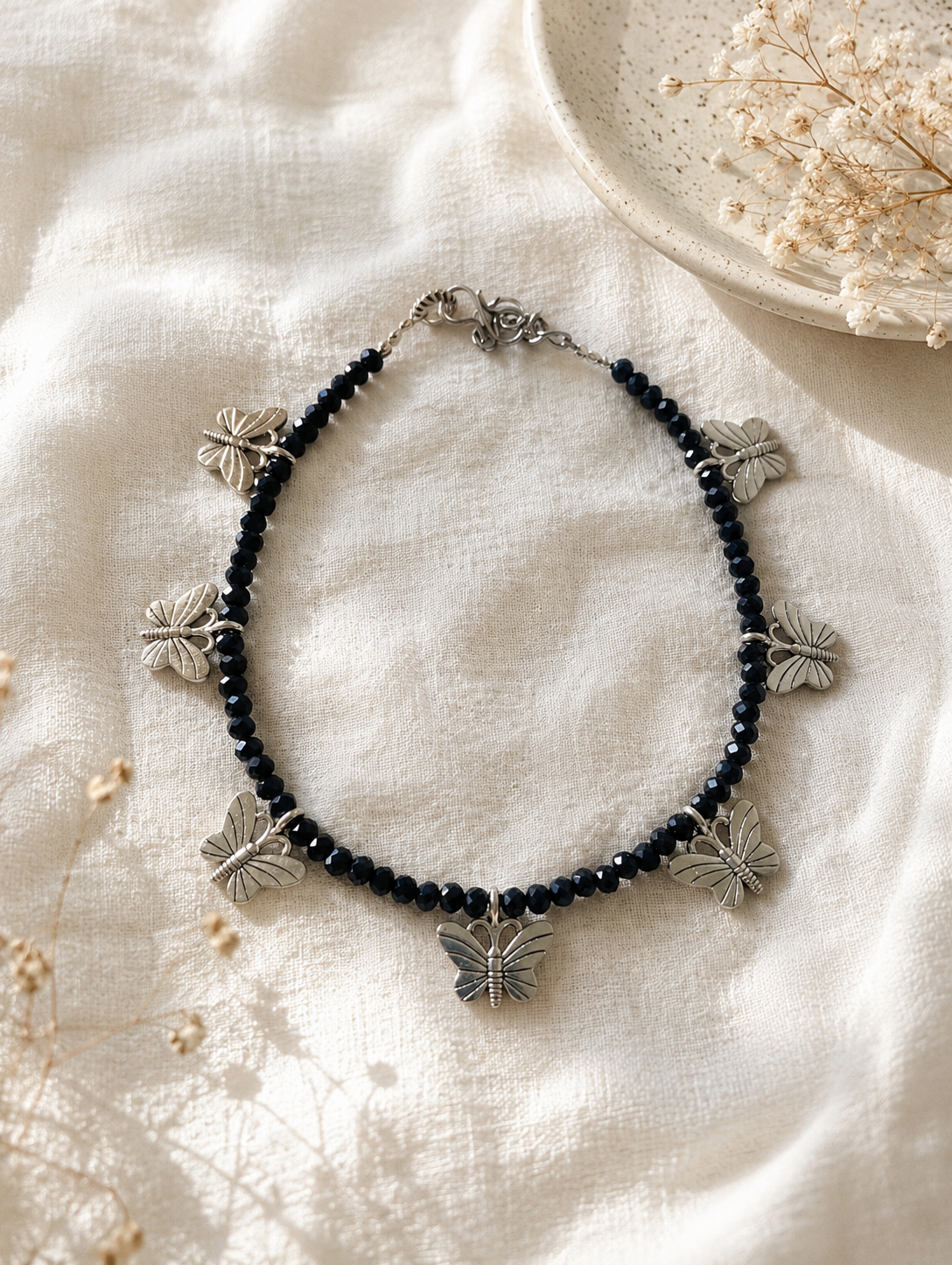 Anklets with black beads and silver butterfly pendants on a textured white surface.