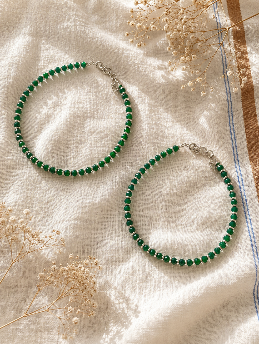 Two green beaded anklet on a textured fabric background with dried flowers.