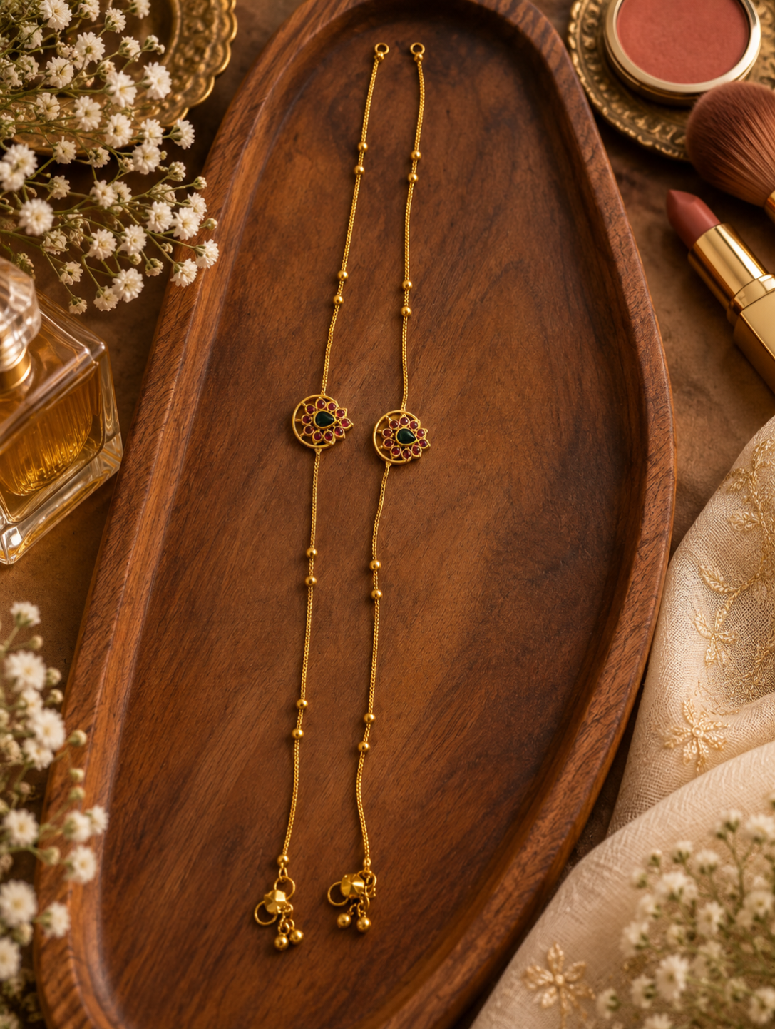 Two gold-toned traditional Indian bangles with circular floral motifs and small green stone accents, displayed on a dark wooden tray with decorative flowers and cosmetics around them.