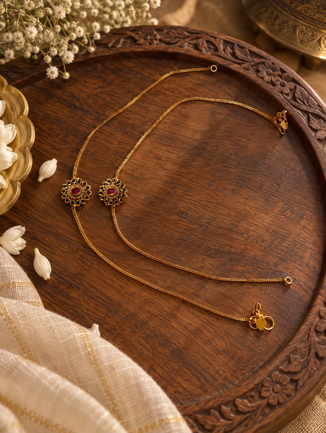 Two gold-toned chain necklaces with ornate floral pendants and red gemstone centers, arranged on a carved wooden tray with decorative flowers and fabric.