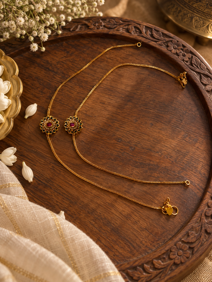 Two gold-toned chain necklaces with ornate floral pendants and red gemstone centers, arranged on a carved wooden tray with decorative flowers and fabric.