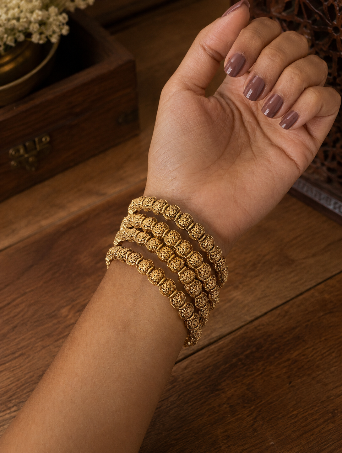 A close-up shot of a person's wrist adorned with a set of four Prafulla traditional gold-plated bangles made of repeating hollow, intricately carved filigree round beads, set against a warm wooden background with a decorative vintage box.