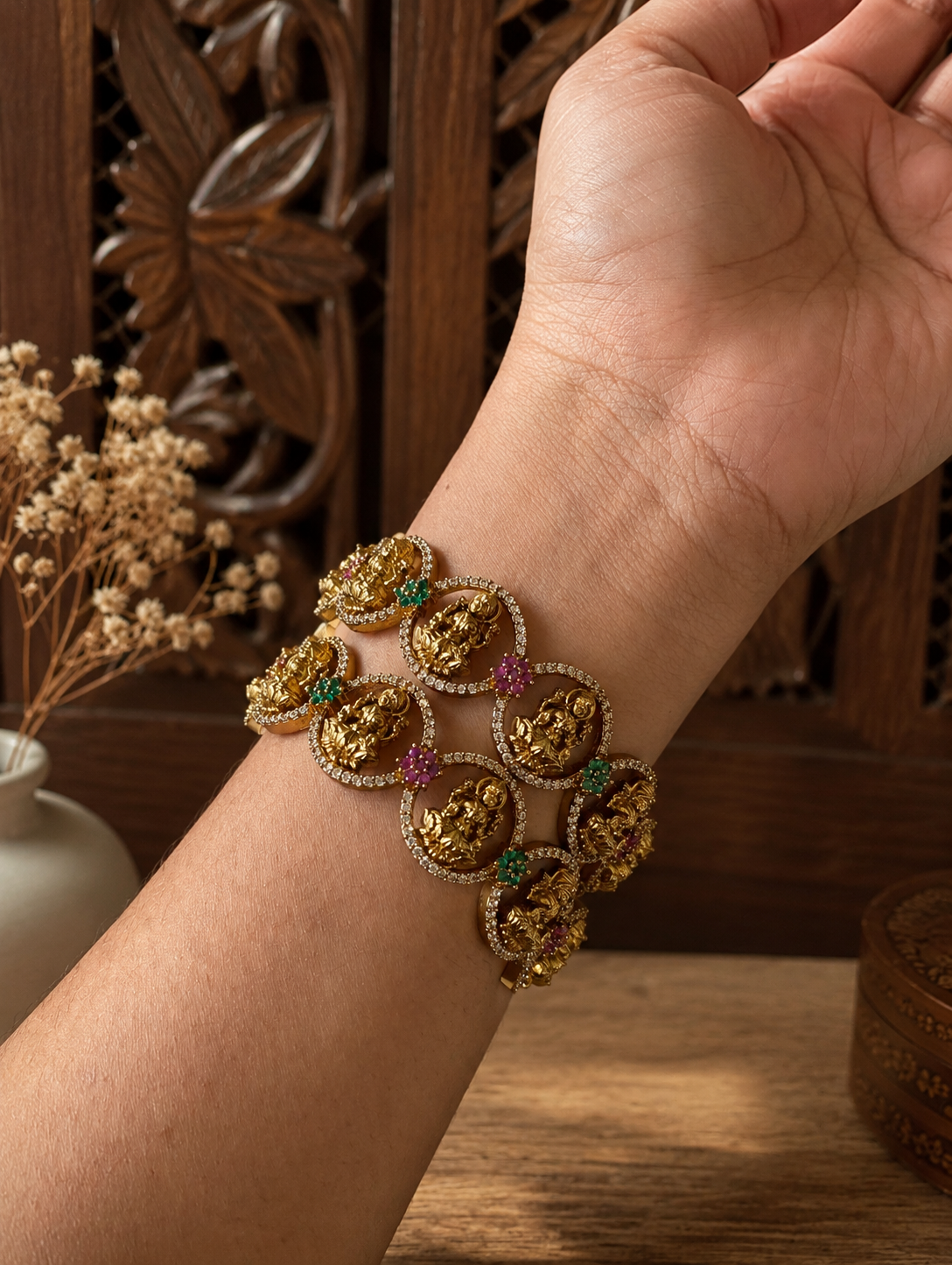 A close-up of a person's wrist wearing the Jhanvi antique gold temple jewelry bracelet, showcasing circular links with detailed Goddess Lakshmi motifs embedded with white CZ, green, and pink stones.