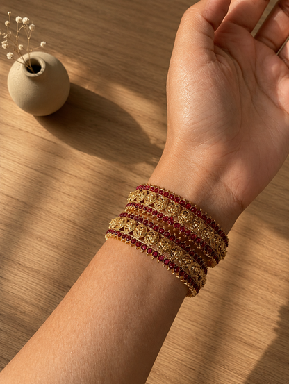 Close-up shot of a person's wrist wearing the Vasantha traditional jewelry set, featuring multiple intricate gold-plated bangles embedded with small, round ruby-red gemstones, set against a warm, sunlit wooden table background with a small ceramic vase.