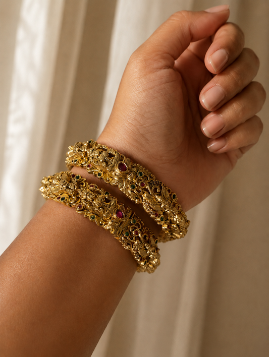 A close-up shot of a model's wrist showcasing a pair of Suneethi traditional heavy gold-plated kada bangles, featuring detailed antique temple-style carvings embedded with small ruby and emerald stones, set against a soft neutral fabric background.