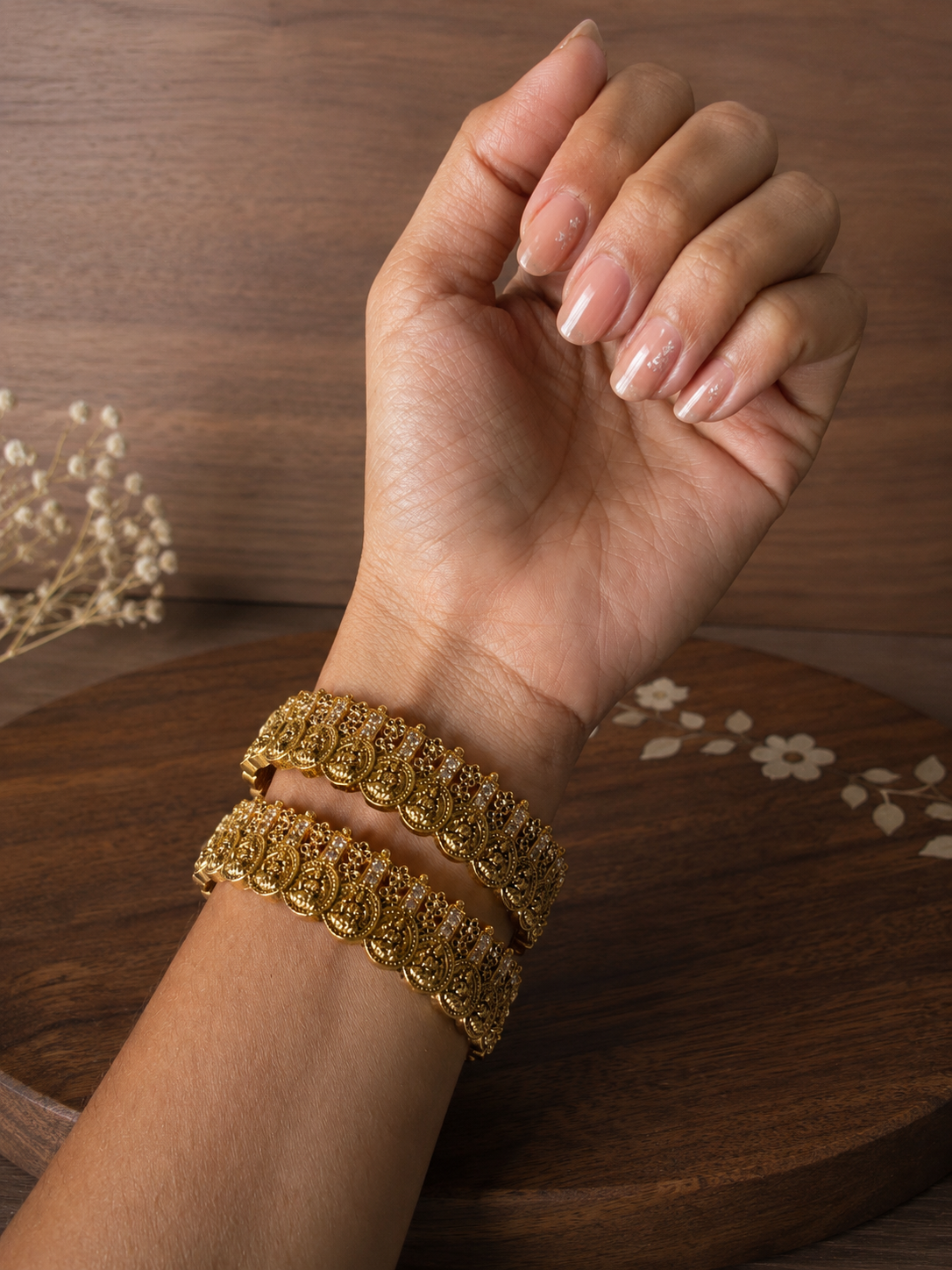 A close-up shot of a person's wrist wearing a pair of Kumudha traditional gold-plated antique bangles featuring repeating embossed Lakshmi coin designs and tiny white zircon stone embellishments, resting against a dark wooden tray background.