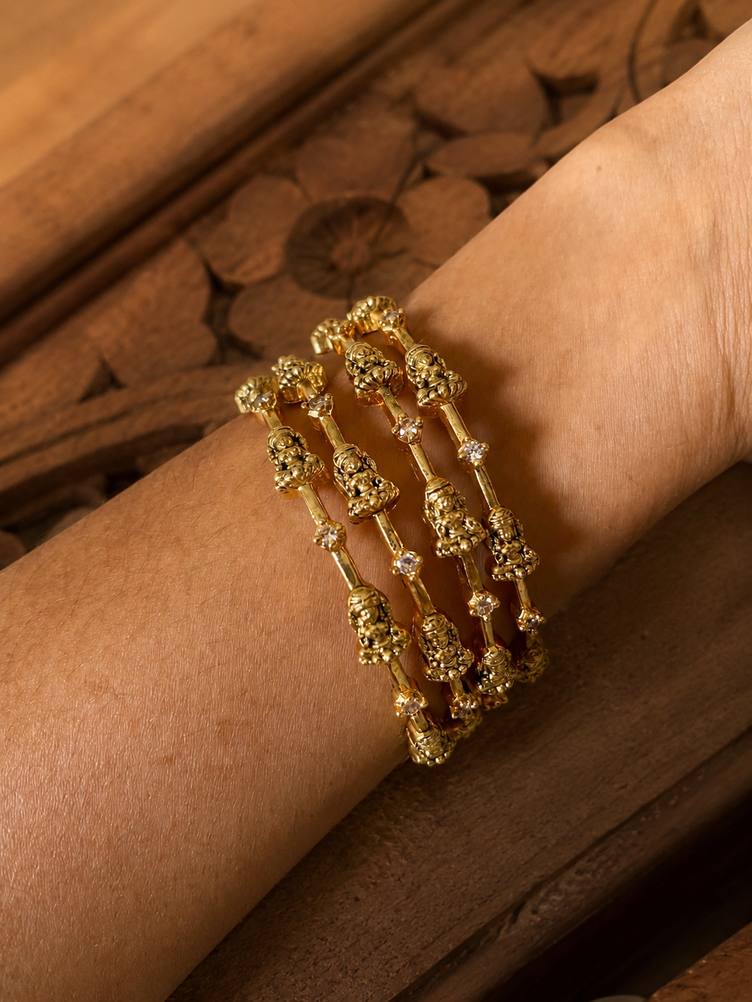 A close-up overhead shot of a person's wrist adorned with a set of four sleek Kanaka gold-plated traditional bangles, featuring repeating miniature embossed Lakshmi idols alternating with tiny, clear white cubic zirconia stones, resting against a dark carved wooden surface.