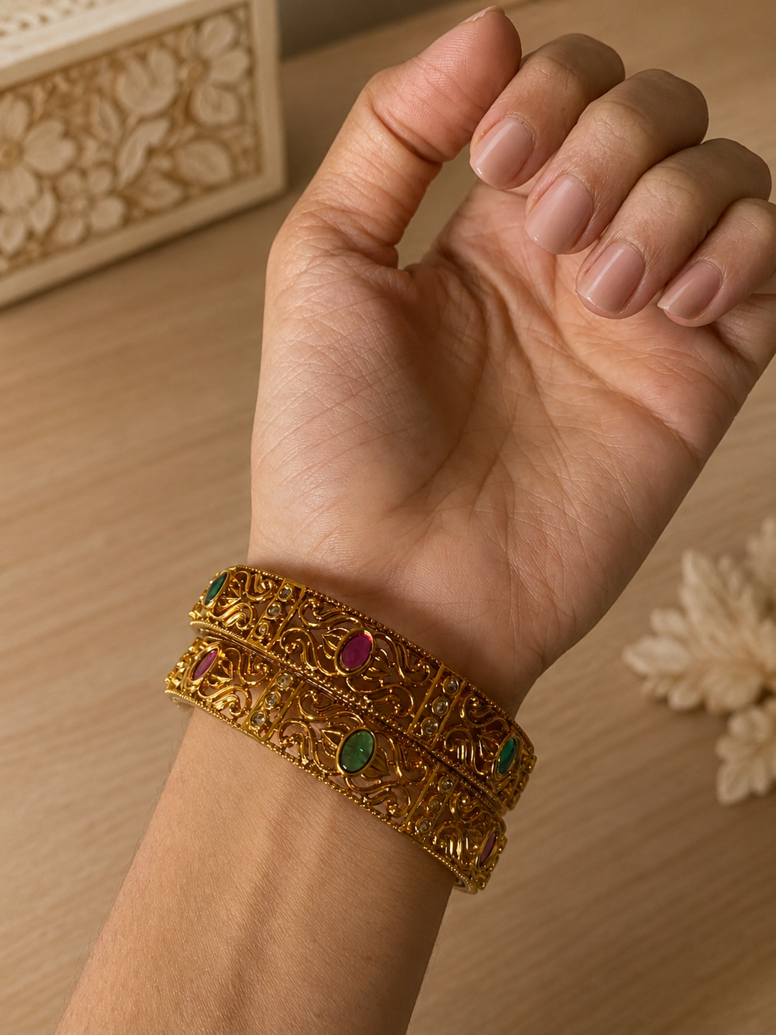 A close-up shot of a model's wrist showcasing a pair of Pranitha traditional gold-plated bangles, designed with intricate openwork filigree scroll patterns and studded with alternating oval ruby, emerald, and small white stones, set against a soft wooden table background.