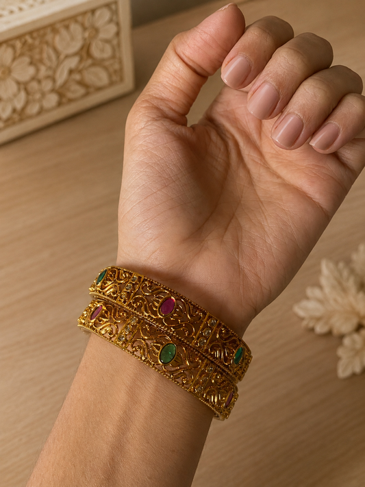 A close-up shot of a model's wrist showcasing a pair of Pranitha traditional gold-plated bangles, designed with intricate openwork filigree scroll patterns and studded with alternating oval ruby, emerald, and small white stones, set against a soft wooden table background.