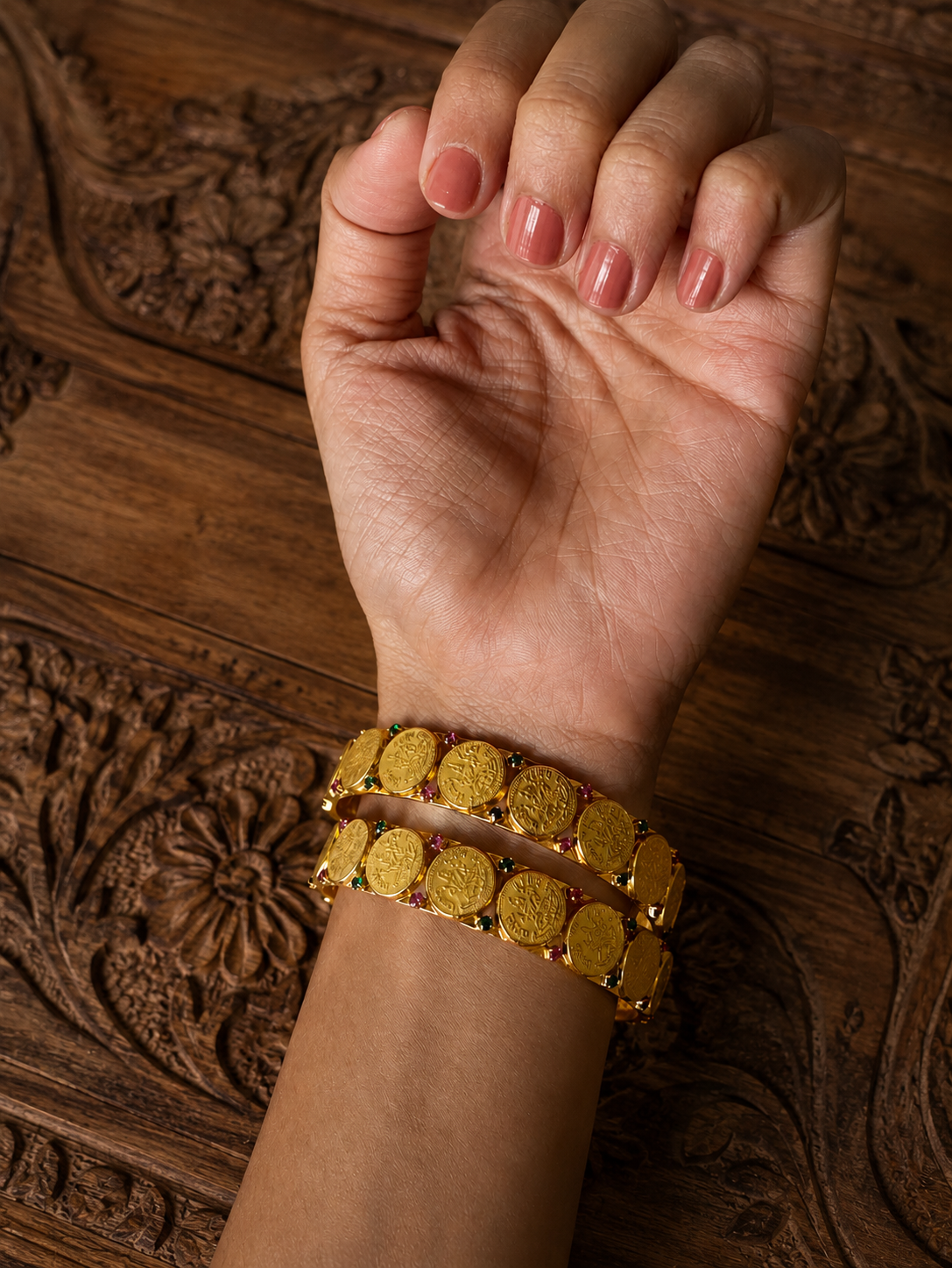 A close-up shot of a person's wrist wearing a pair of Kasu traditional gold-plated antique bangles, composed of overlapping Lakshmi coin motifs separated by tiny pink ruby and green emerald stones, set against a dark carved wooden background.