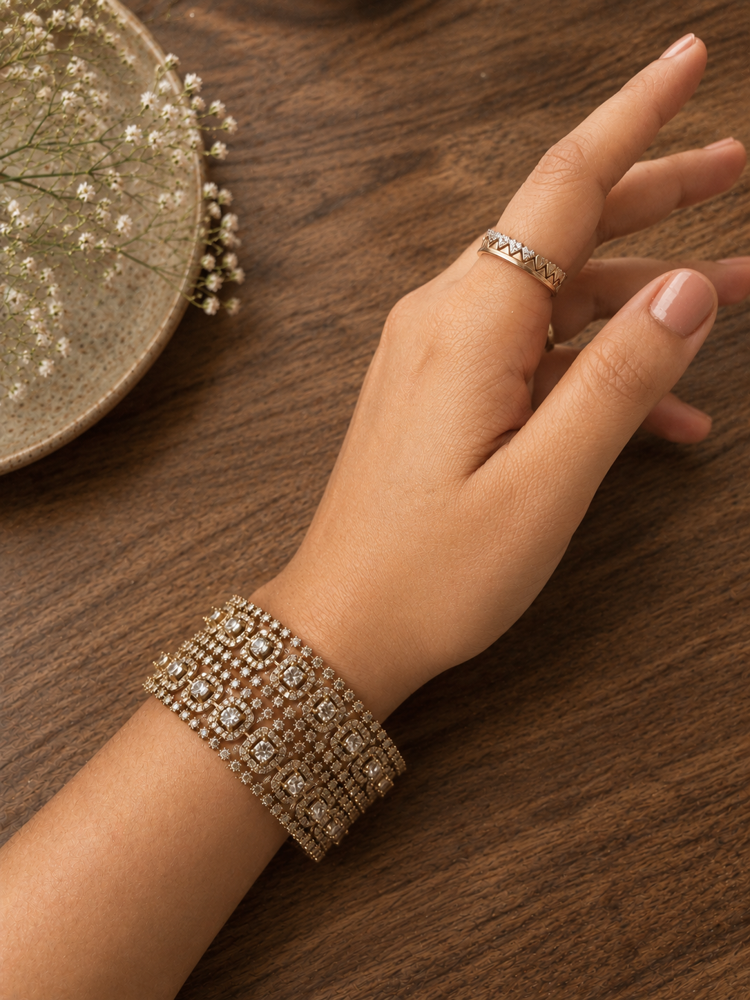 A top-down shot of a person's wrist featuring the Bhramari broad, multi-layered gold-plated bracelet encrusted with rows of brilliant square and round white CZ stones, styled alongside a matching geometric ring on a wooden table background.