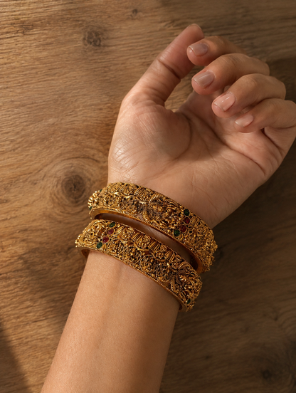 A high-angle close-up shot of a person's wrist wearing a pair of thick, antique gold-plated kada bangles featuring highly detailed traditional Nakshi temple-style carvings and tiny ruby and emerald embellishments, resting against a rustic wooden background.