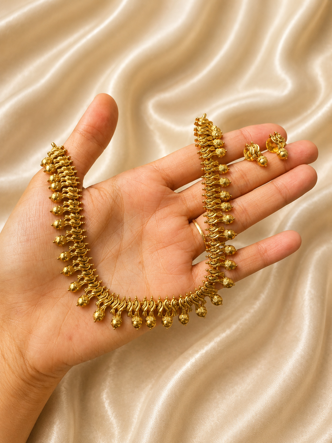 A traditional South Indian antique gold-plated short necklace and matching tiny stud earrings held on a hand against a champagne satin fabric, featuring a closely knit row of wavy gold links finished with small hanging golden metal balls.