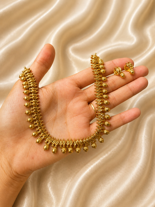A traditional South Indian antique gold-plated short necklace and matching tiny stud earrings held on a hand against a champagne satin fabric, featuring a closely knit row of wavy gold links finished with small hanging golden metal balls.